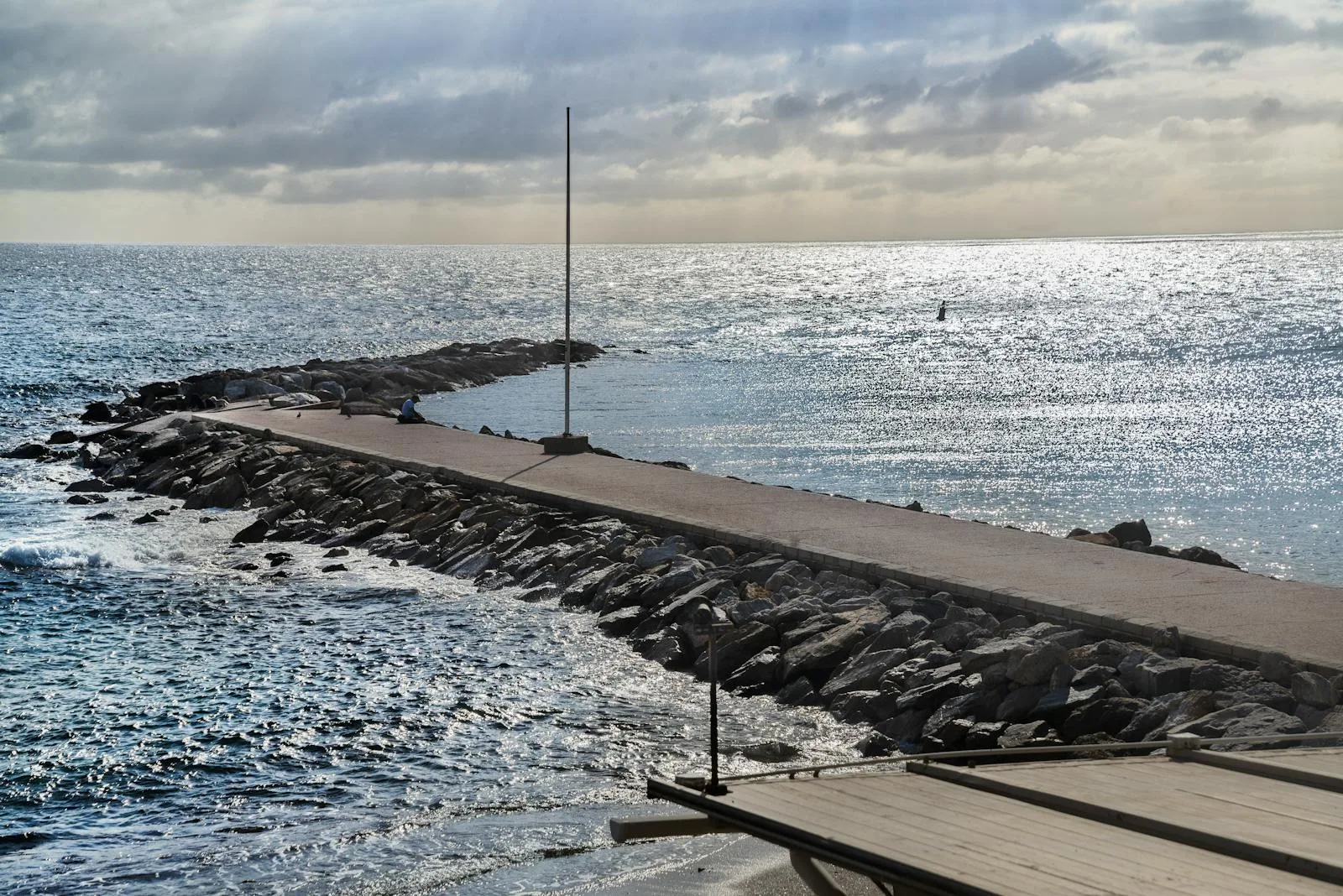 Calm beach scene with a paved jetty extending into the ocean under a cloudy sky.